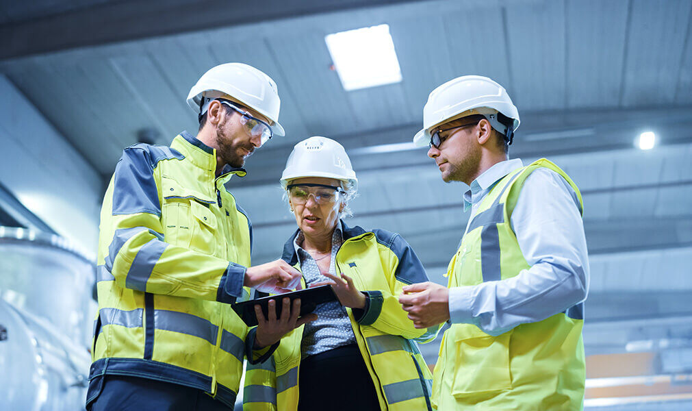 Two men and a woman with PPE discuss a document in factory floor